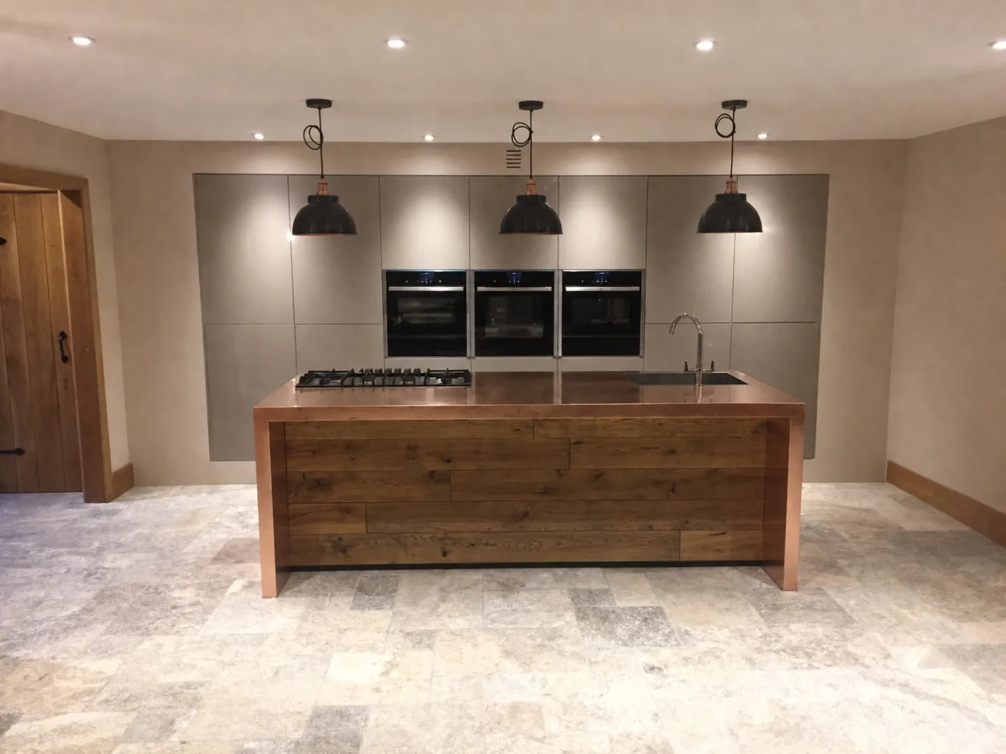 Bespoke kitchen island clad in reclaimed oak with a copper worktop, set against handleless grey cabinetry and a travertine stone floor
