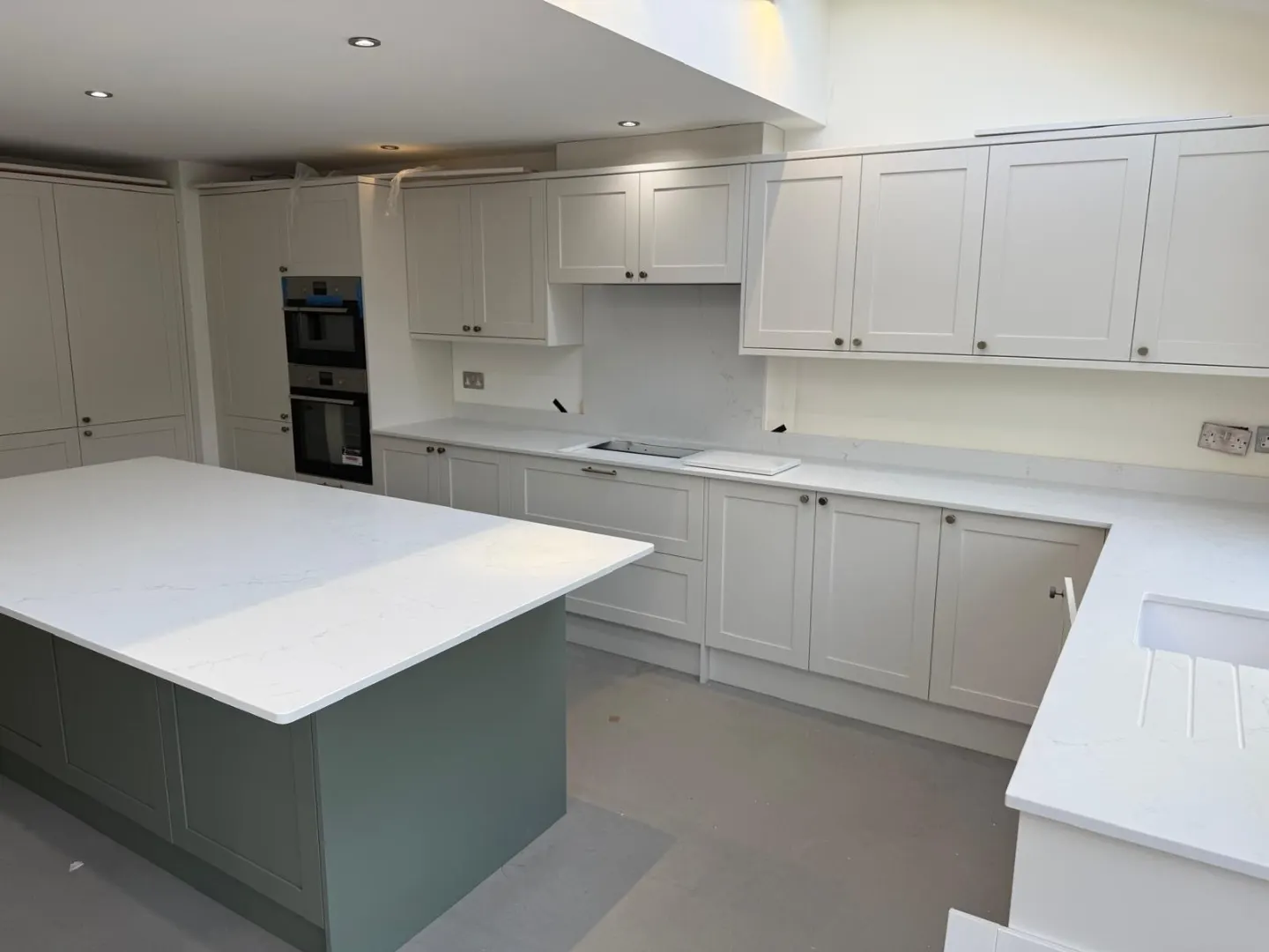 Newly installed pale grey shaker kitchen with quartz worktops, a sage green island and integrated double oven, photographed before flooring was laid